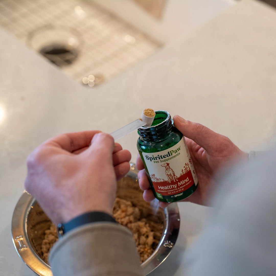 Person opening a bottle of Healthy Mind supplement with a pet's food bowl in the background.